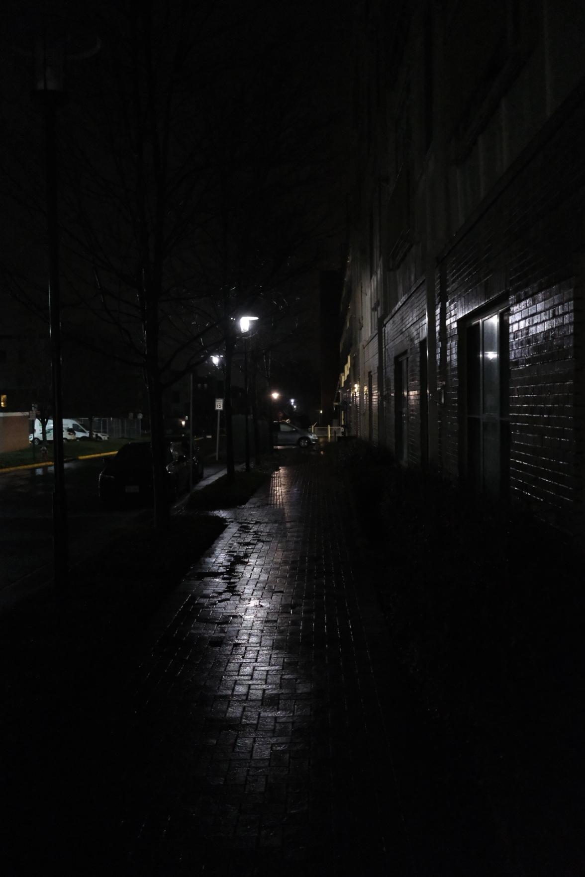 Staring down the long brick sidewalk, after a rain storm left everything a bit wet. Next to the path is a brick wall that is also wet. Both are illuminated by a lamp posts lining the path, causing some start streaks of light along their otherwise dead of the night feel.
